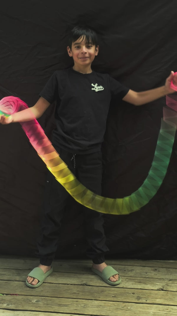 Young boy against a dark backdrop demonstrating springbending or slinky manipulation tricks with a stretched Rainbow HyperSpring plastic spring toy