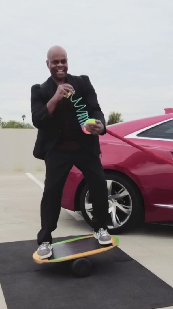 Man in a suit balancing on a balance board outdoors next to a red car while demonstrating springbending or slinky manipulation tricks with a Rainbow HyperSpring plastic spring toy